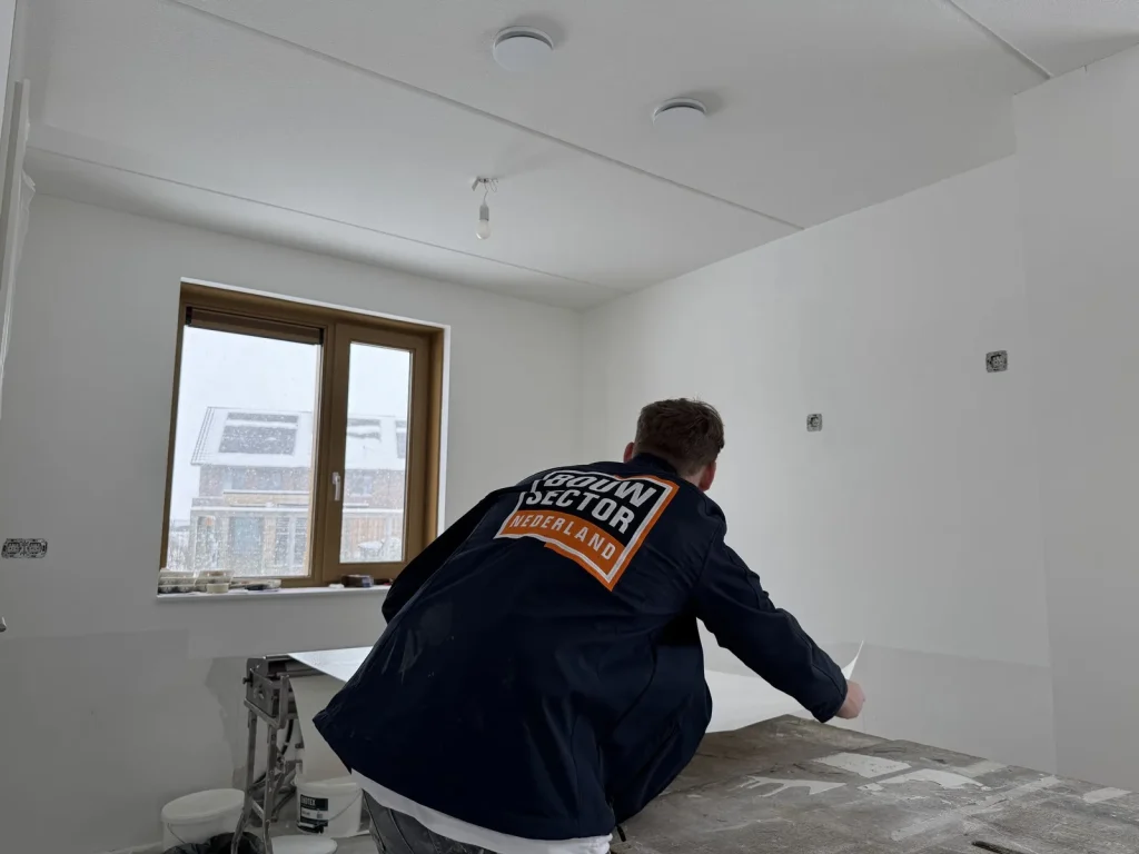 Behanger in donkere werkjas brengt vlies aan op de wand van een net opgeleverde woning